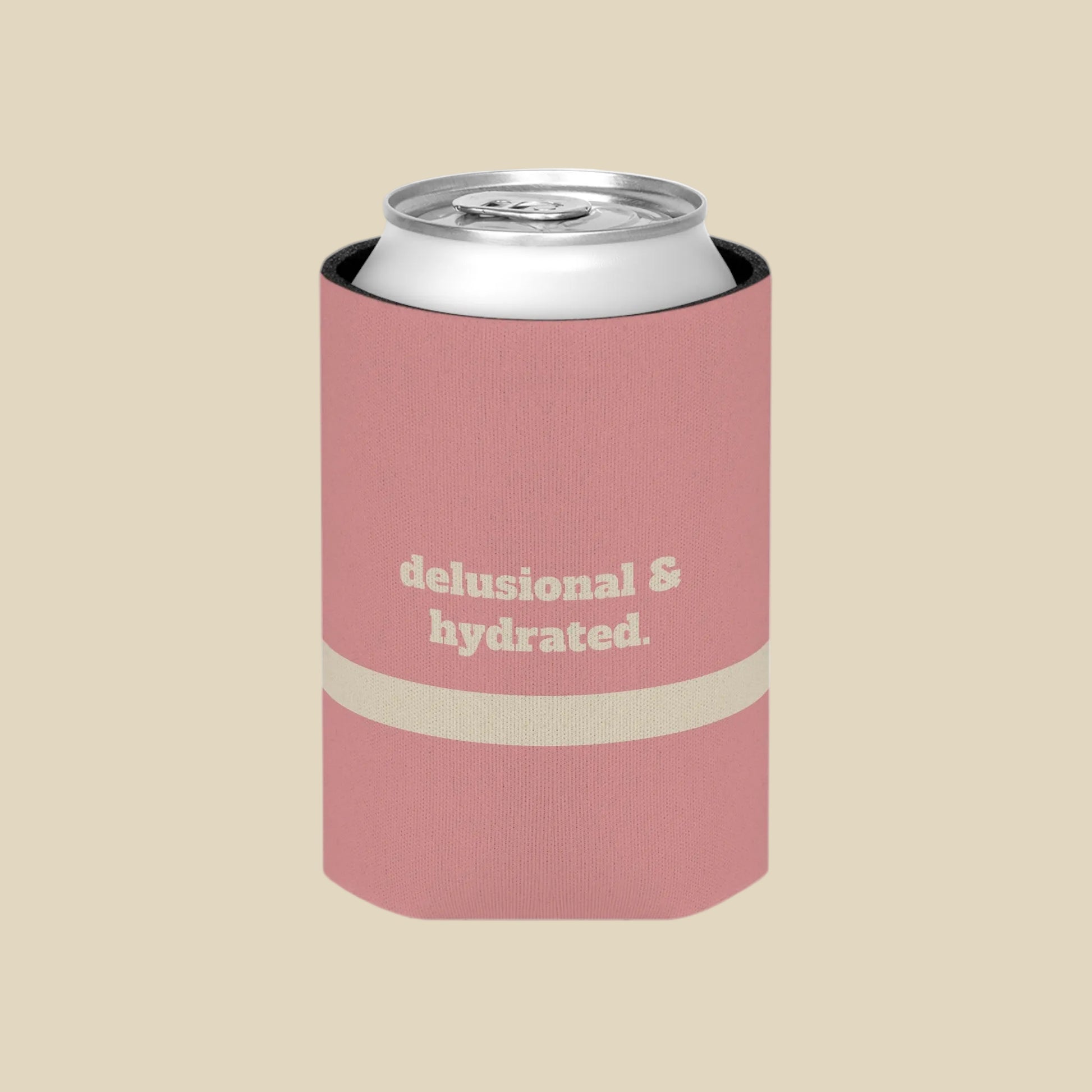 A pink can cooler sleeve holding a silver aluminum can, featuring the text 'delusional & hydrated.' in beige on the front, set against a plain white background.