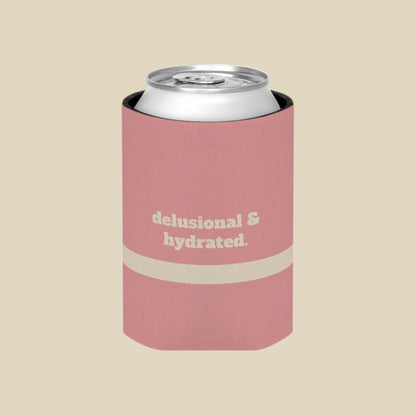 A pink can cooler sleeve holding a silver aluminum can, featuring the text 'delusional & hydrated.' in beige on the front, set against a plain white background.