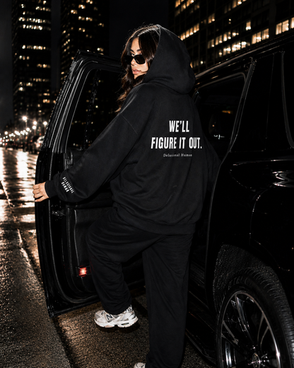 Person wearing a black hoodie with text standing next to a car at night.