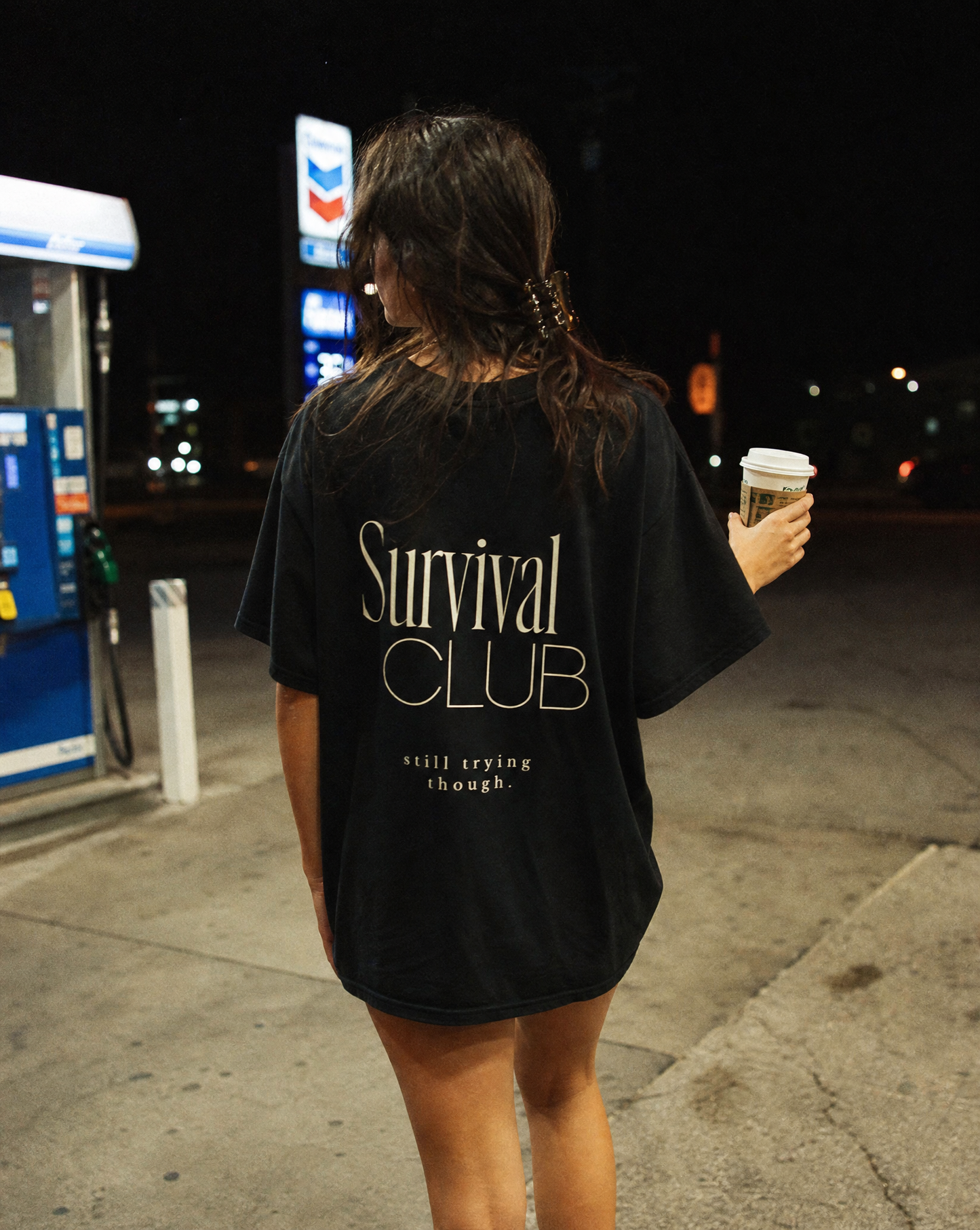 Person wearing a black 'Survival Club' t-shirt holding a coffee cup at night.