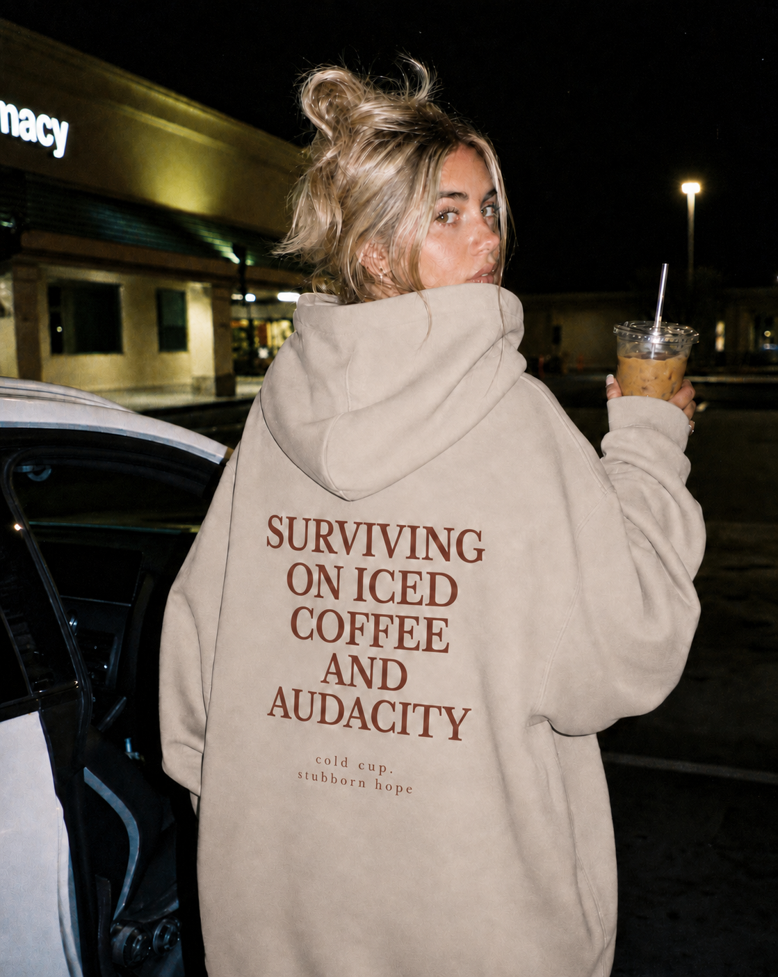 Person wearing a hoodie with text, holding an iced coffee, in a parking lot at night.