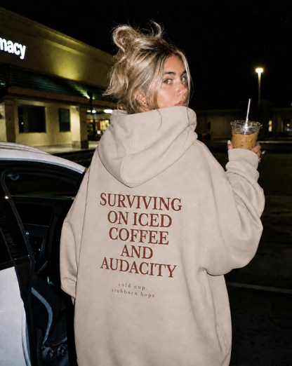 Person wearing a hoodie with text, holding an iced coffee, in a parking lot at night.