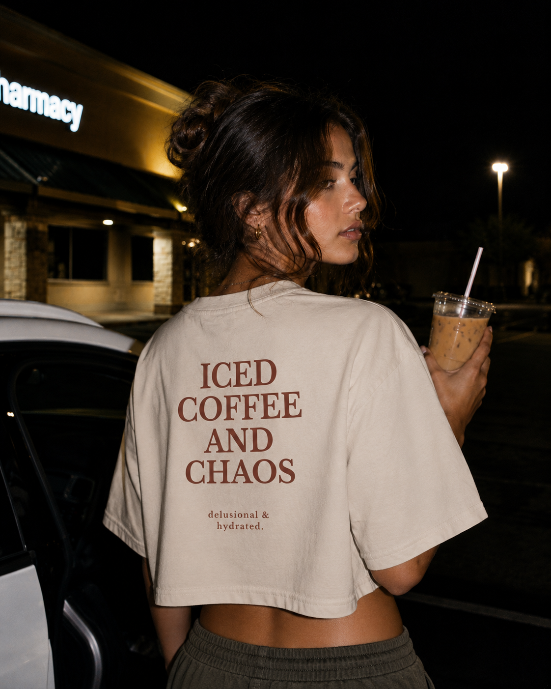 Person wearing a beige t-shirt with 'ICED COFFEE AND CHAOS' text, holding an iced coffee drink at night.