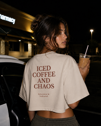 Person wearing a beige t-shirt with 'ICED COFFEE AND CHAOS' text, holding an iced coffee drink at night.