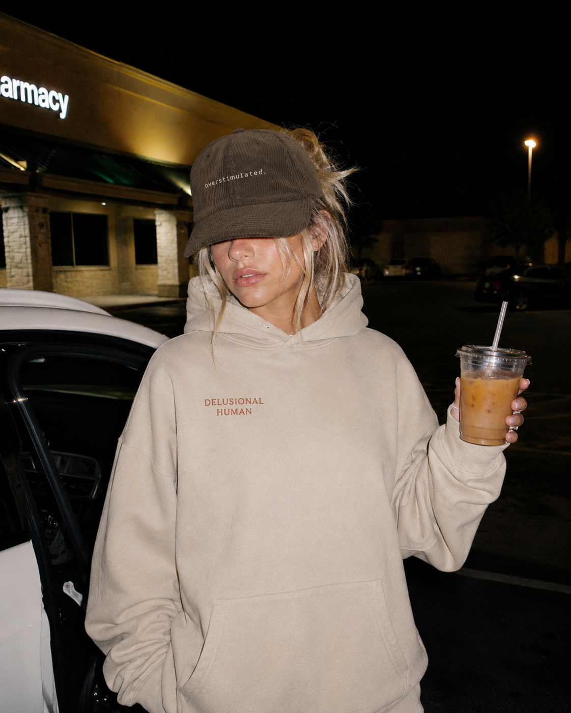 Person wearing a beige hoodie and dark cap, holding a drink at night.